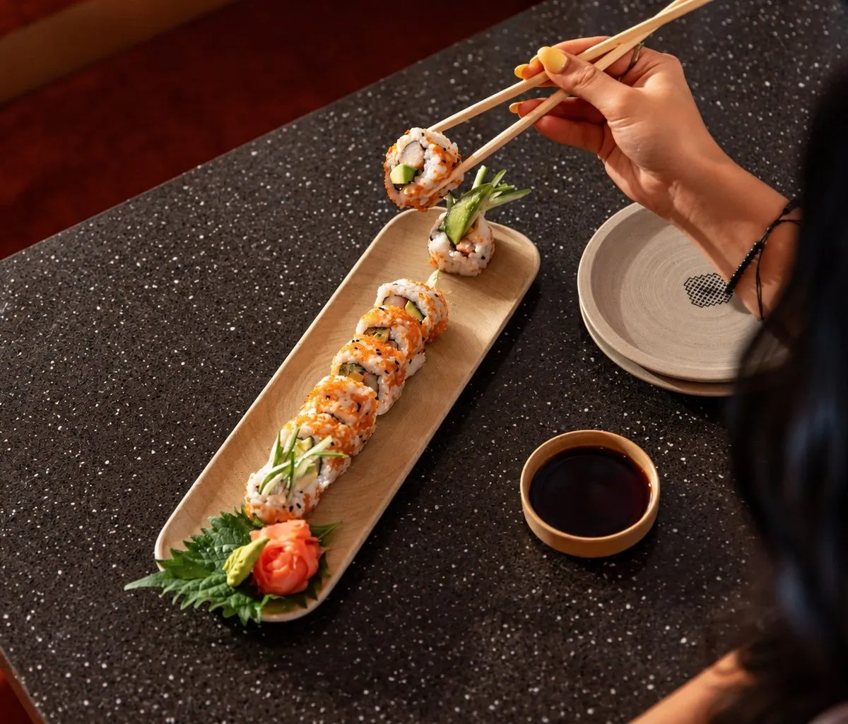 A board of sushi rolls and a dish of soy on a dark counter